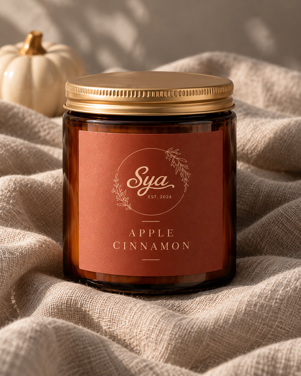 Vela Sya en frasco ámbar con tapa dorada y etiqueta color terracota Apple Cinnamon, sobre tela beige iluminada con luz cálida.