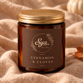 Cinnamon & Clove candle in an amber glass jar with a gold lid, resting on beige fabric.