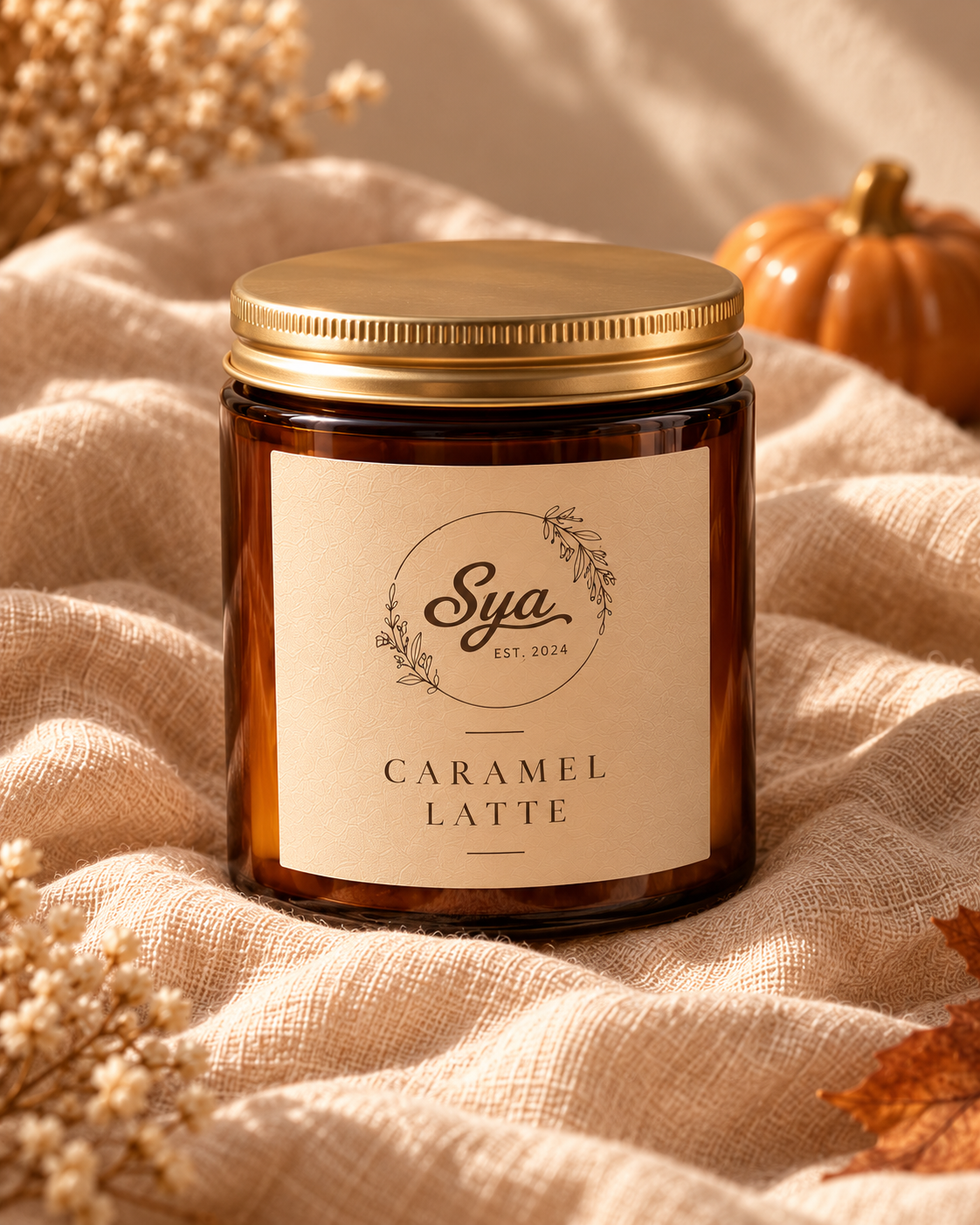 Vela Sya en frasco ámbar con tapa dorada y etiqueta crema Caramel Latte, sobre tela beige con flores secas y decoración otoñal.