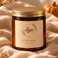 Caramel Latte candle in an amber glass jar with a gold lid, resting on beige fabric.